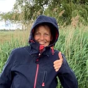 A woman in a dark blue raincoat with pink piping stands in a field smiling and showing a thumbs up