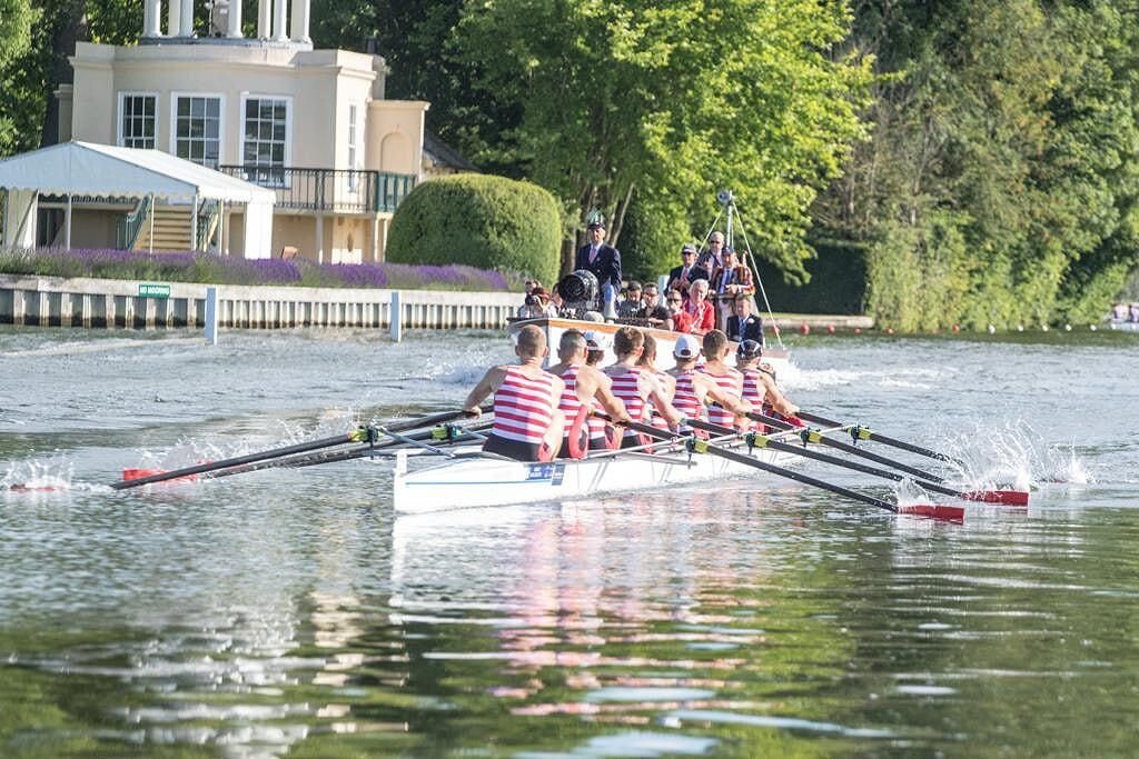 A 8+ rows down the Henley Royal Regatta race course. The rowers have red oars and are wearing red and white striped clothing. The boat is followed by an umpire's launch. It's a bright and sunny day.