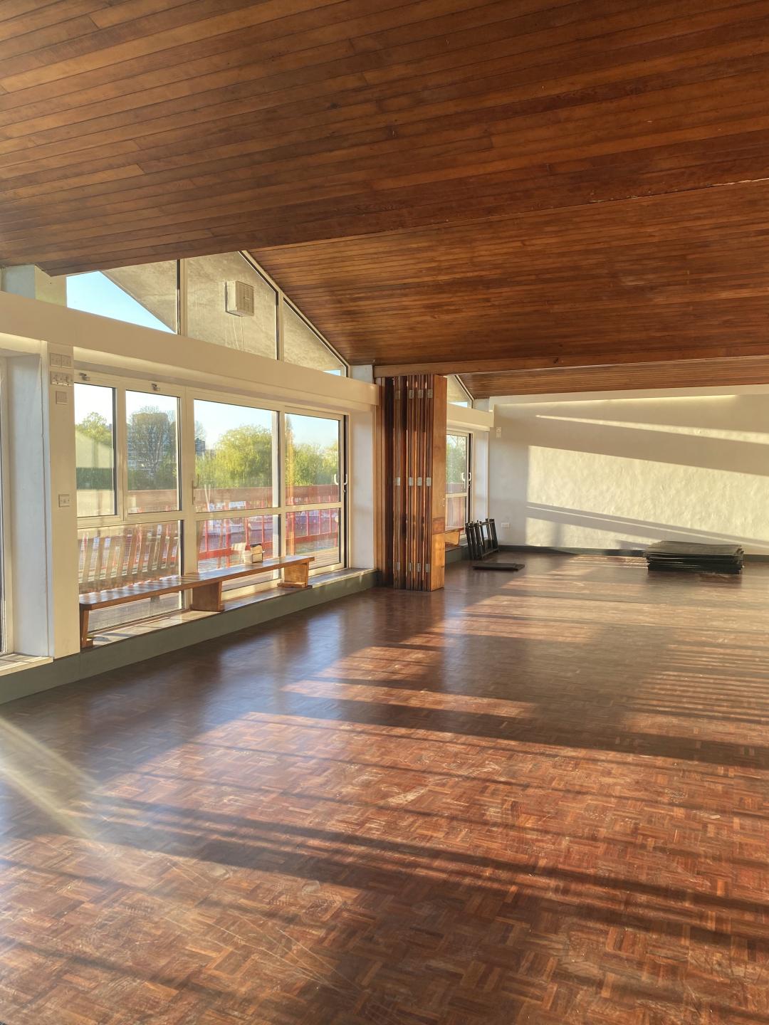 A hall with wooden floors and wooden ceiling. On the left it has floor to ceiling windows with a view of the river thames.