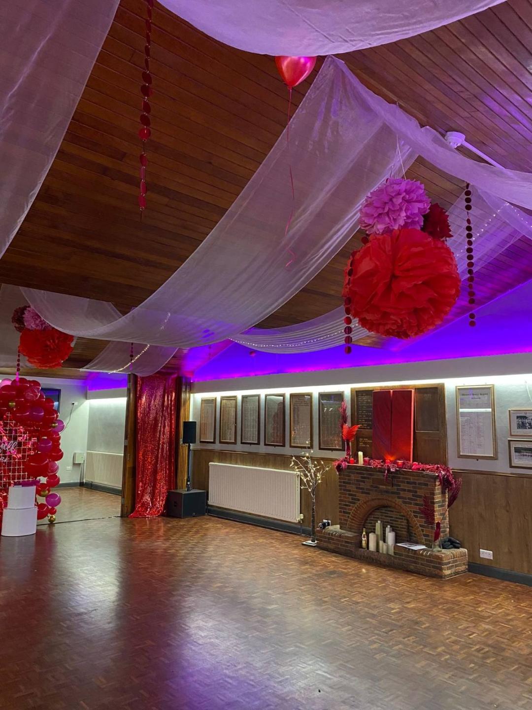 A hall with wooden floor and ceilings draped with white fabric and red decorations. There is a brick fireplace against the wall.
