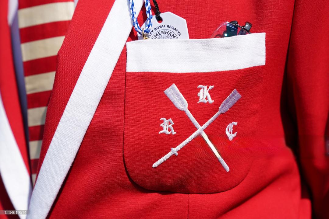 A red Kingston Rowing Club blazer showing a pair of white crossed oars with the letters "K", "R", and "C" written around them. The pocket contains an entry badge for Remenham Club and a pair of glasses. Also visible is a red and white horizontal striped tie.
